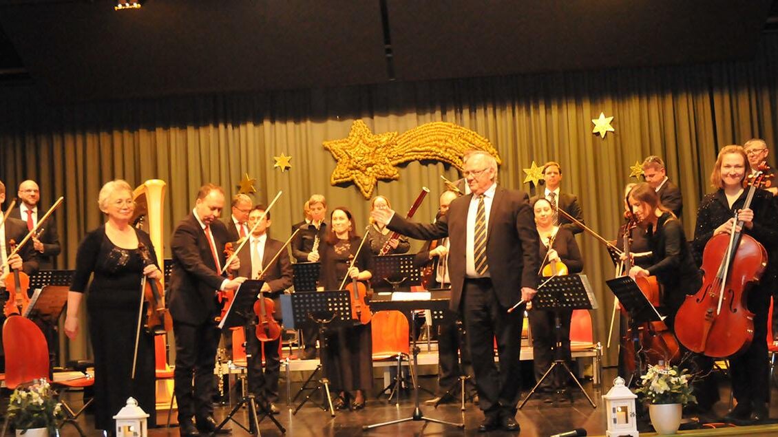 Ein Orchester steht auf der Bühne und applaudiert. Im Hintergrund sind dekorative Sterne und Stühle sichtbar. | © Siegel Sabine