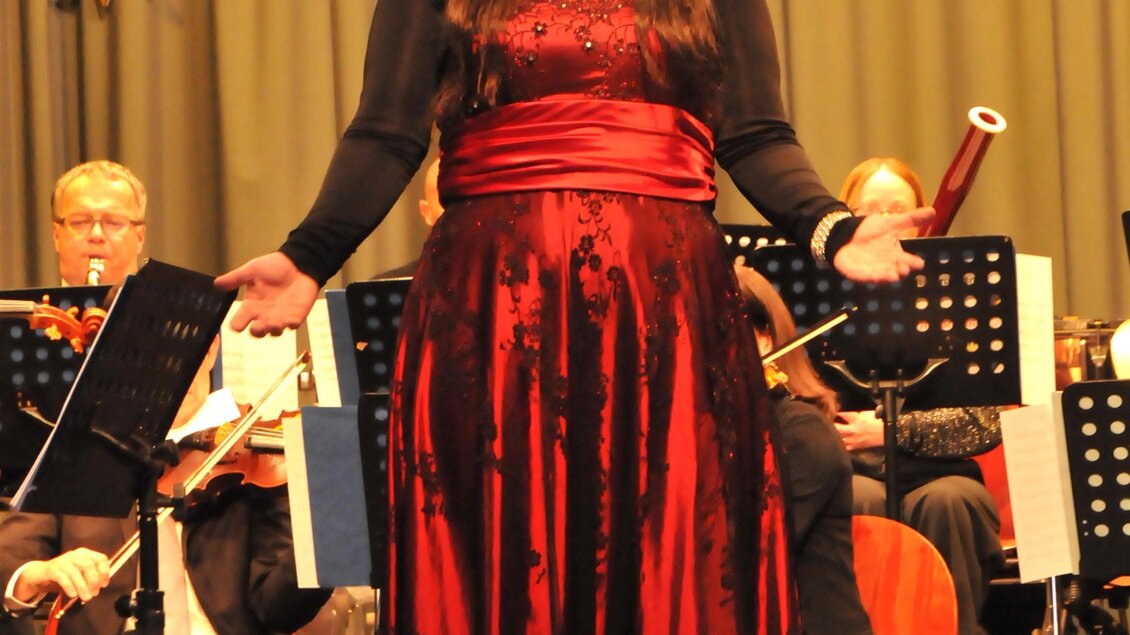 Eine Frau in einem roten Abendkleid singt auf der Bühne. Im Hintergrund spielt ein Orchester. | © Siegel Sabine