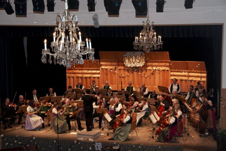 Ein Orchester spielt auf einer Bühne in festlicher Atmosphäre. Anwesend sind Musiker in traditioneller Kleidung, umgeben von eleganten Kristalllüstern. | © Stadtgemeinde Bad Aussee