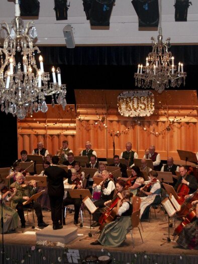 An orchestra is playing on a stage in a festive atmosphere. Present are musicians in traditional clothing, surrounded by elegant crystal chandeliers. | © Stadtgemeinde Bad Aussee