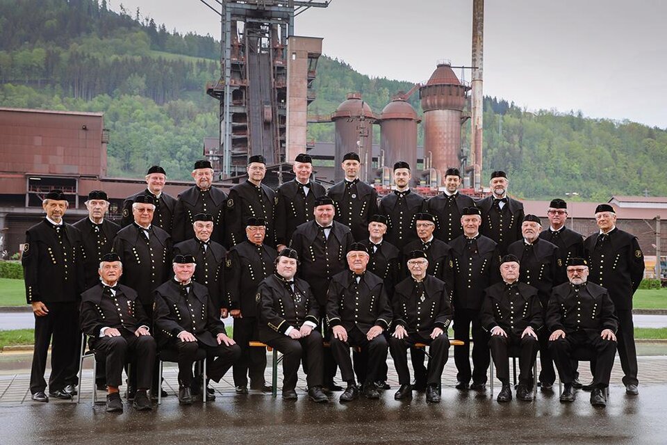 Eine Gruppe von Männern in traditionellen Hüten und Kleidung steht in einem alten Industriegebiet. Im Hintergrund sind große, historische Anlagen und grüne Hügel zu sehen. | © Werkschor Donawitz