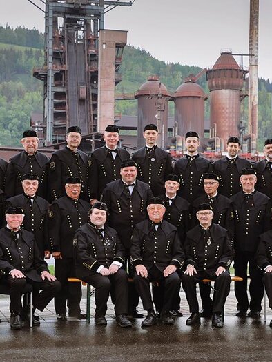 A group of men in traditional hats and clothing stands in an old industrial area. In the background, large historic facilities and green hills can be seen. | © Werkschor Donawitz