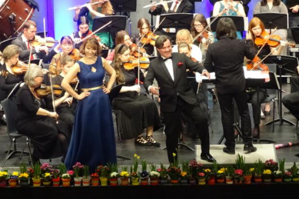 Eine Aufführung mit einem Orchester auf der Bühne. Zwei Personen, elegant gekleidet, präsentieren das Konzert, während das Orchester im Hintergrund spielt. | © KK