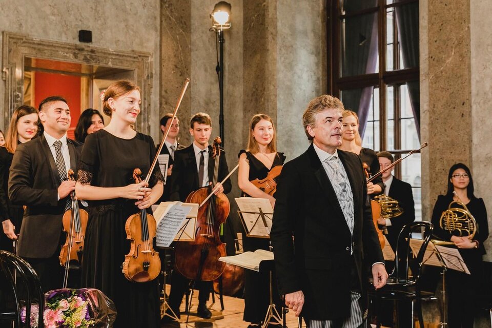 Ein Orchester steht auf der Bühne, die Musiker lächeln und applaudieren. Im Vordergrund steht der Dirigent, der das Publikum anblickt. | © TvMurau