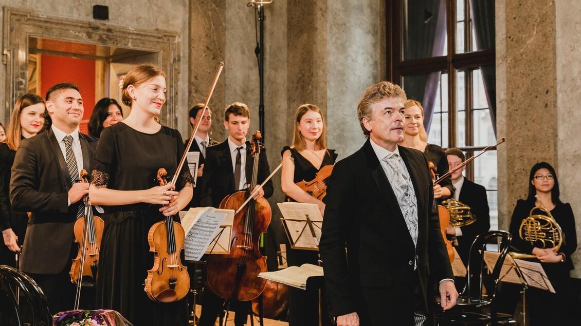 Ein Orchester steht auf der Bühne, die Musiker lächeln und applaudieren. Im Vordergrund steht der Dirigent, der das Publikum anblickt. | © TvMurau