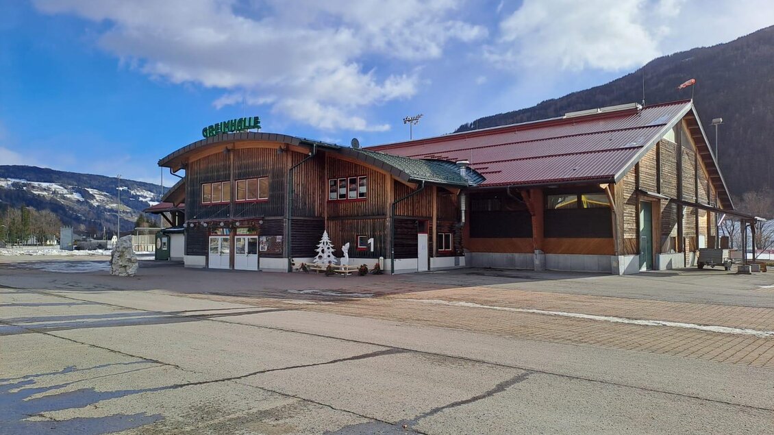 Ein großes, modernes Gebäude mit Holzfassade und rotem Dach. Es ist ein klarer Tag mit einigen Wolken und einer schönen Berglandschaft im Hintergrund. | © Tourismusverband Murau