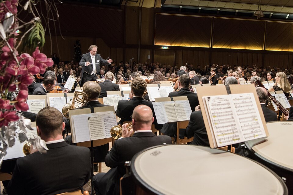 Ein Orchester spielt auf einer Bühne mit einem Dirigenten. Das Publikum sitzt im Vordergrund und hört aufmerksam zu. | © Johann Zugschwert