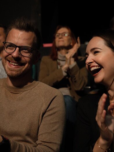 A couple smiles and applauds in a crowd of spectators. The atmosphere is cheerful and relaxed. | ©  achromaticphotography