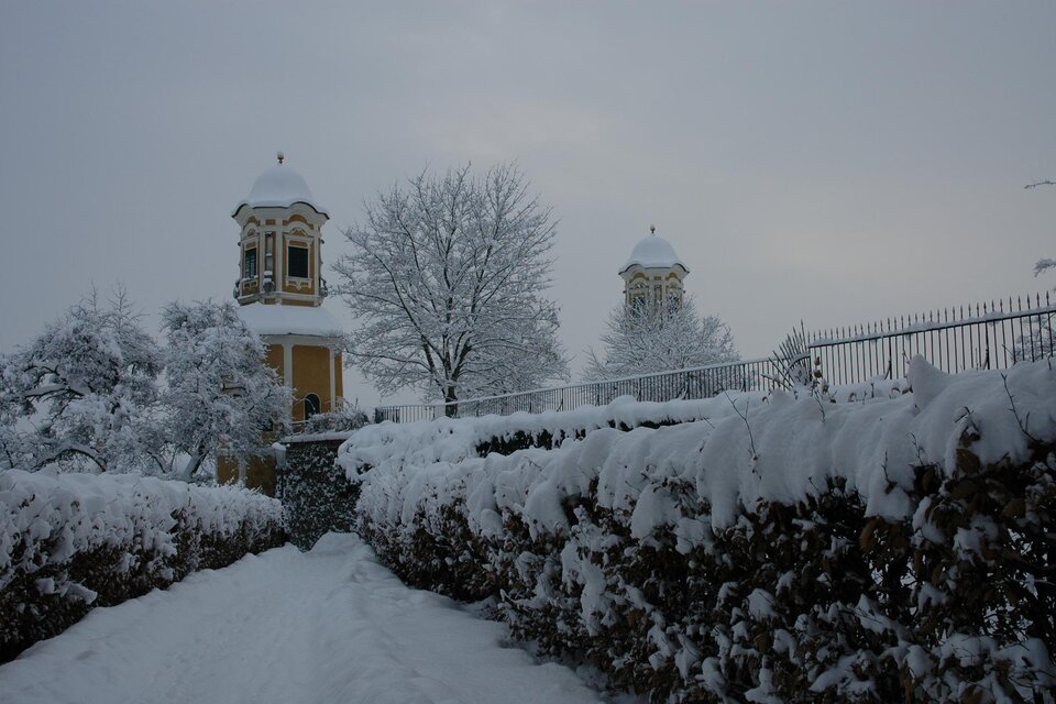 Eine verschneite Landschaft mit hohen Hecken und zwei Türmen im Hintergrund. Die Bäume sind ebenfalls mit Schnee bedeckt und der Himmel ist grau. | © Riffel Helmuth