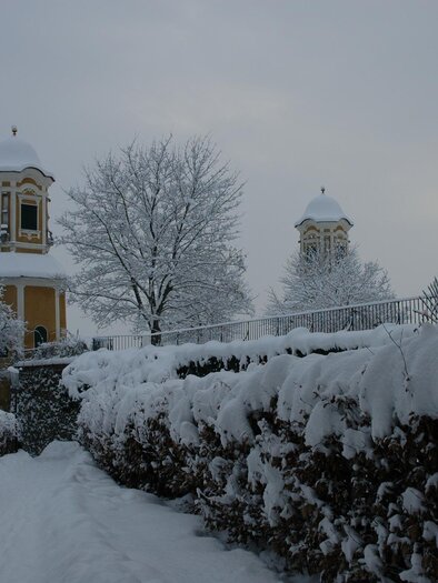 A snowy landscape with tall hedges and two towers in the background. The trees are also covered in snow, and the sky is gray. | © Riffel Helmuth