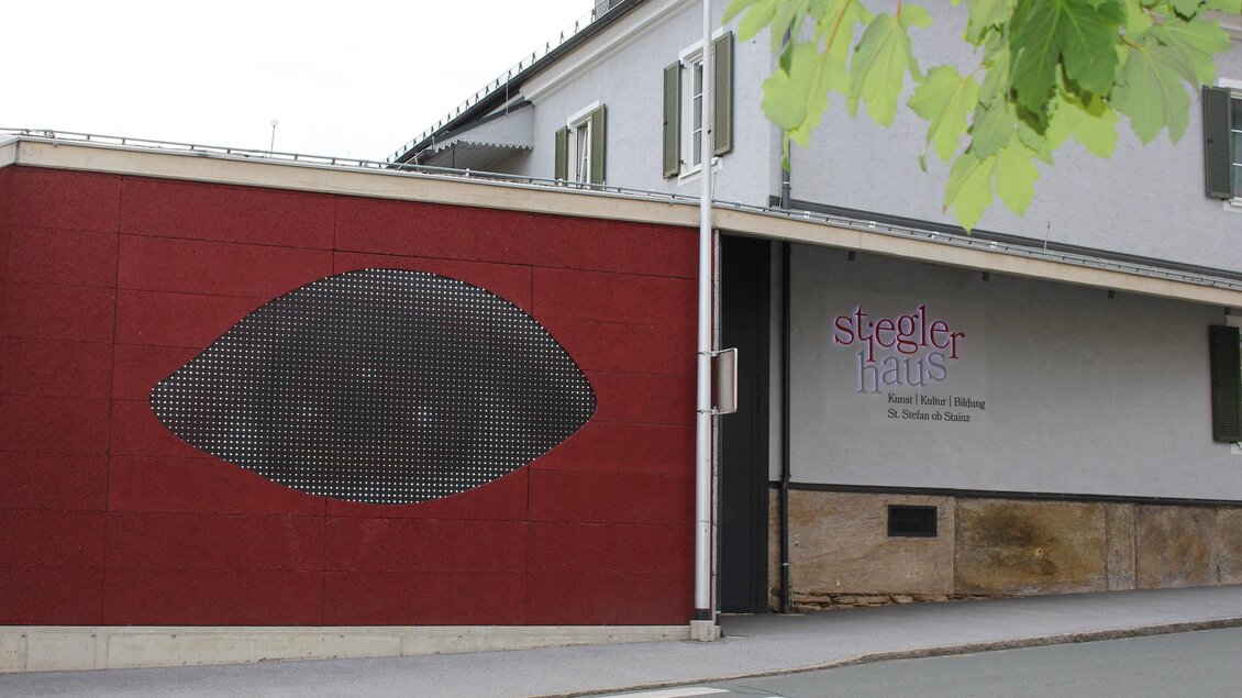 Ein modernes Gebäude mit einer auffälligen roten Wand, die ein stilisiertes Auge zeigt. Daneben befindet sich das Schild "Stegler Haus". | © TV Südsteiermark - Irene Löschnig