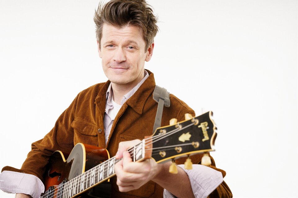 Portrait eines Mannes mit halbakustischer Gitarre vor hellem, neutralem Hintergrund; er trägt eine braune Cordjacke und blickt mit leichtem Lächeln in die Kamera. | © Stefanos Notopoulos