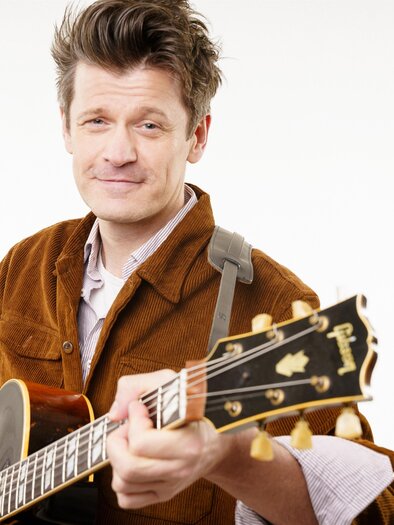 Portrait eines Mannes mit halbakustischer Gitarre vor hellem, neutralem Hintergrund; er trägt eine braune Cordjacke und blickt mit leichtem Lächeln in die Kamera. | © Stefanos Notopoulos