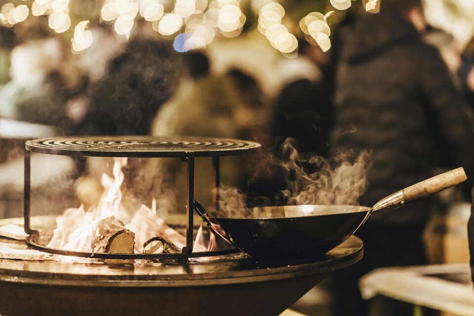 Ein Grill mit einer Pfanne auf dem Feuer, umgeben von verschwommenen Lichtern und Menschen. Es herrscht eine gemütliche Stimmung. | © Kunsthandwerk Schloss Burgau