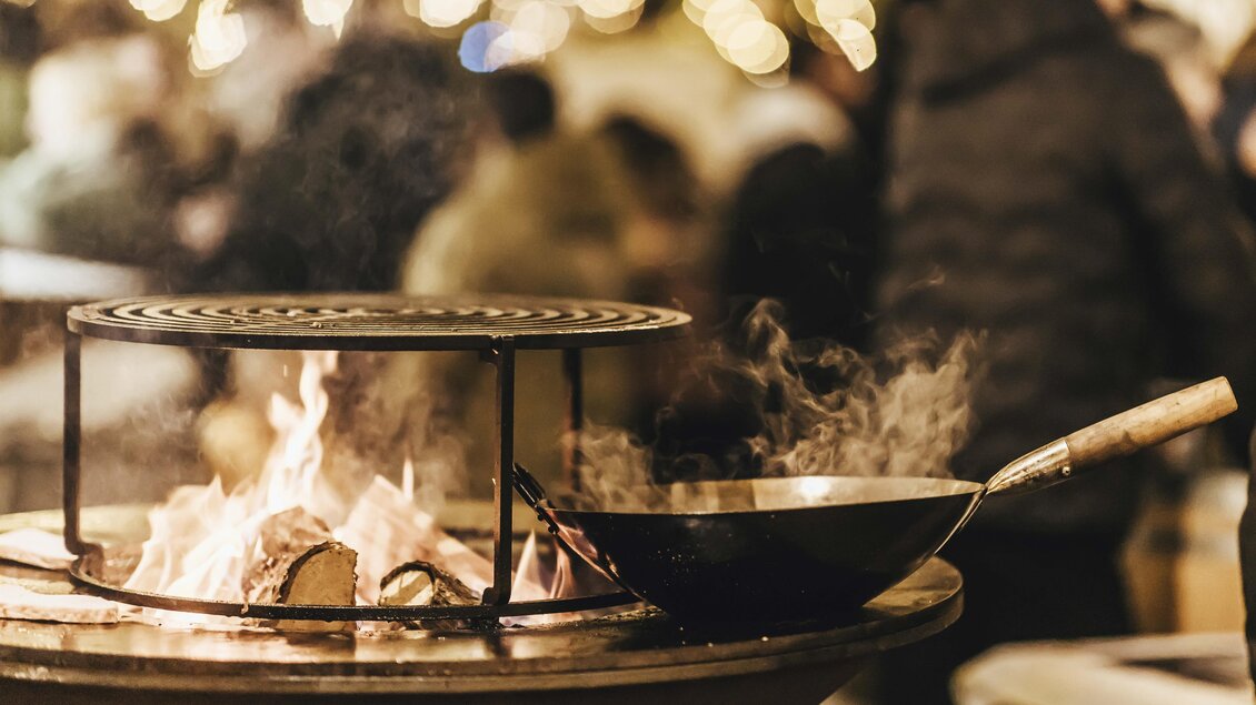 Ein Grill mit einer Pfanne auf dem Feuer, umgeben von verschwommenen Lichtern und Menschen. Es herrscht eine gemütliche Stimmung. | © Kunsthandwerk Schloss Burgau