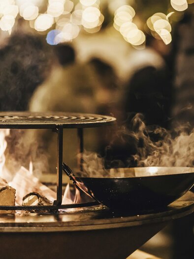 A grill with a pan over the fire, surrounded by blurred lights and people. There is a cozy atmosphere. | © Kunsthandwerk Schloss Burgau