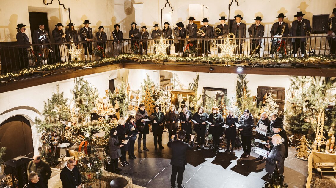 Ein Chorgesang in einem festlich dekorierten Raum mit vielen Weihnachtsbäumen. Die Sänger stehen auf einem Balkon und sind elegant gekleidet. | © Kunsthandwerk Schloss Burgau