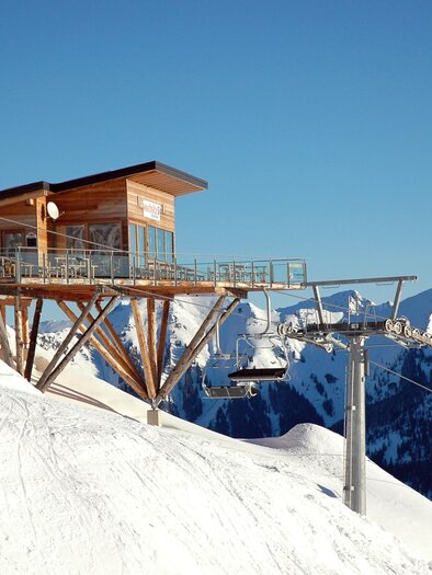 A modern cabin on a snowy mountain. In the background, majestic mountains and a clear blue sky can be seen. | © Riesneralm Bergbahnen/Erwin Petz