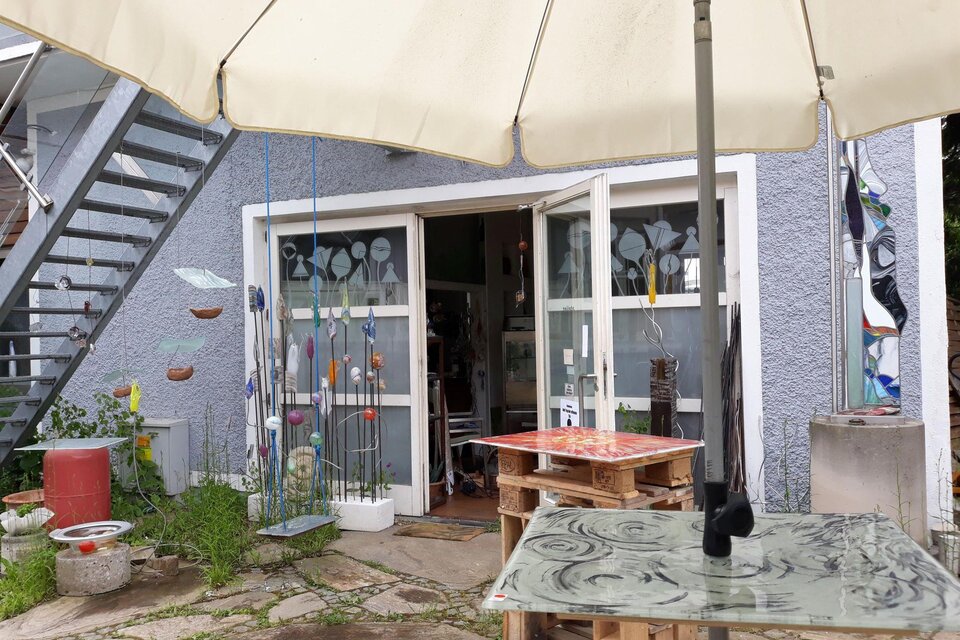 Ein gemütlicher Außenbereich mit Tischen aus Glas und einem Sonnenschirm. Im Hintergrund befindet sich ein Eingang zu einem Atelier oder einer Galerie. | © TV Suedsteiermark - Grete Faßwald