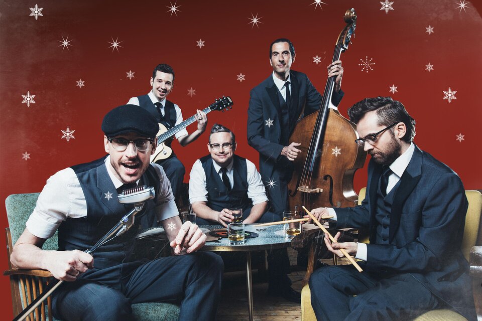 Eine Band mit fünf Musikern in eleganter Kleidung spielt in einem festlichen Ambiente. Der Hintergrund ist rot mit Schneeflocken, und sie scheinen Spaß zu haben. | © Marija Kanizaj