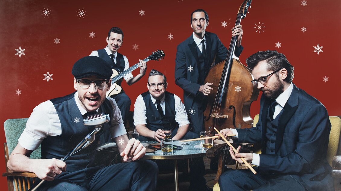 Eine Band mit fünf Musikern in eleganter Kleidung spielt in einem festlichen Ambiente. Der Hintergrund ist rot mit Schneeflocken, und sie scheinen Spaß zu haben. | © Marija Kanizaj