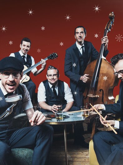 A band with five musicians in elegant clothing is playing in a festive atmosphere. The background is red with snowflakes, and they seem to be having fun. | © Marija Kanizaj