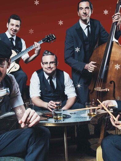 A band with five musicians in elegant clothing sits around a table. The background is red with white snowflakes.