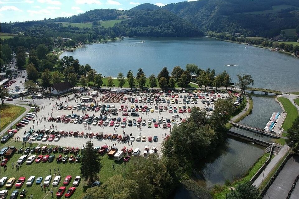 Eine große Versammlung von Autos in einem Parkgebiet am Wasser. Im Hintergrund sind grüne Hügel und ein blauer Himmel zu sehen. | © Werner Thaler