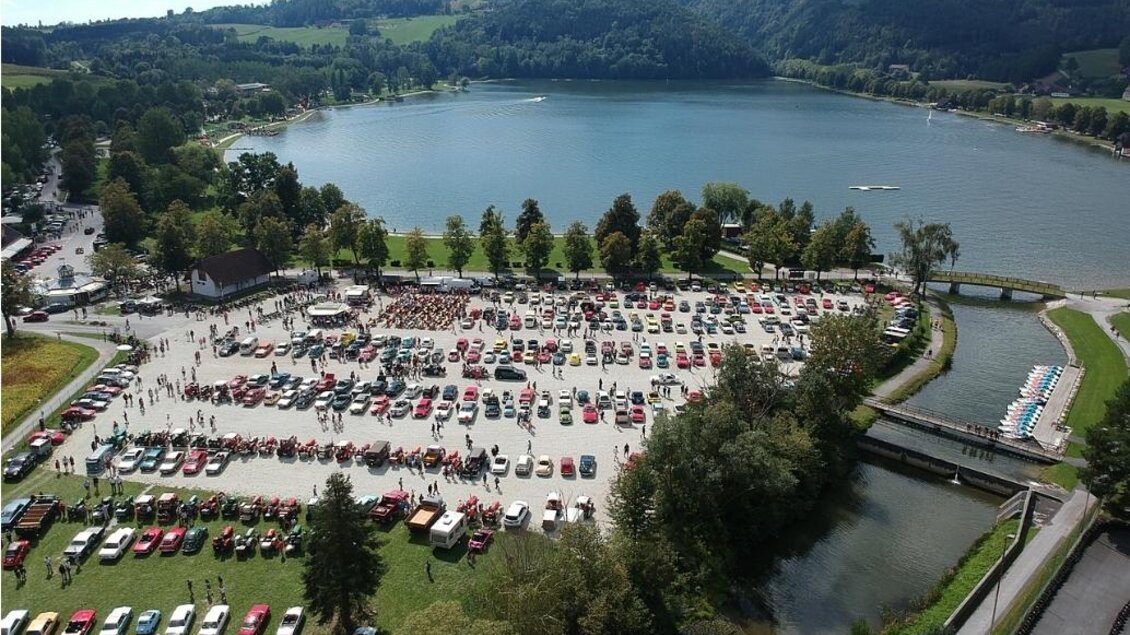 Eine große Versammlung von Autos in einem Parkgebiet am Wasser. Im Hintergrund sind grüne Hügel und ein blauer Himmel zu sehen. | © Werner Thaler
