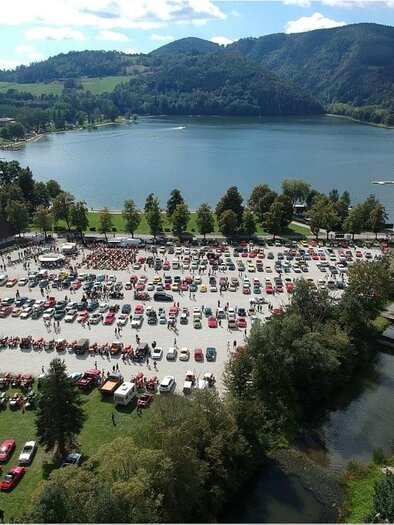 A large gathering of cars in a park area by the water. In the background, green hills and a blue sky are visible. | © Werner Thaler