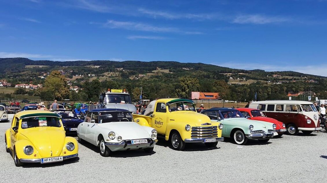 Eine Reihe klassischer Autos steht auf einem Parkplatz. Im Hintergrund sind grüne Hügel und ein blauer Himmel zu sehen. | © Werner Thaler