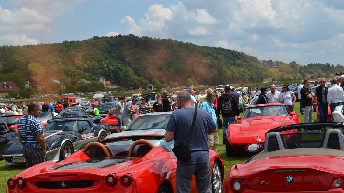 Eine lebendige Automobilmesse mit vielen roten Sportwagen und einer großen Menschenmenge. Im Hintergrund sind grüne Hügel und ein blauer Himmel zu sehen. | © SSW
