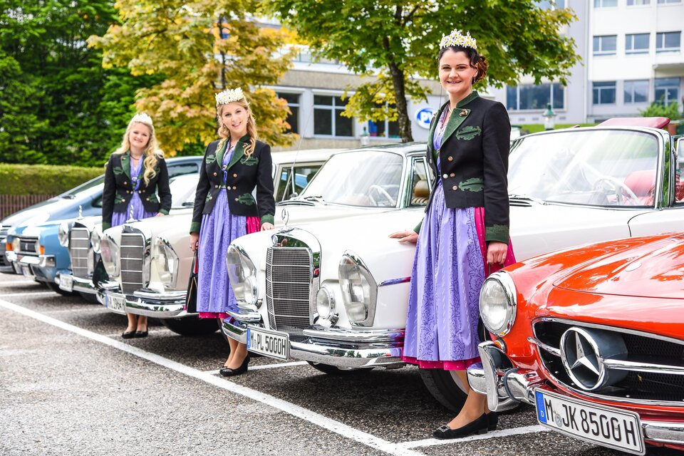 Drei Frauen in traditionellen Kleidern stehen neben vintage Autos. Der Parkplatz ist von Bäumen umgeben und es herrscht eine festliche Atmosphäre. | © Herbert Sams
