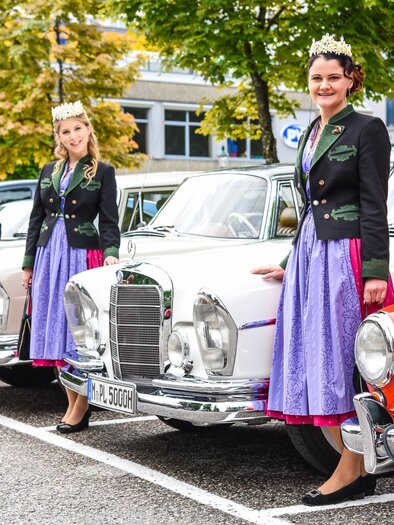 Three women in traditional attire stand next to a row of classic cars. In the background, trees and a building are visible. | © Herbert Sams