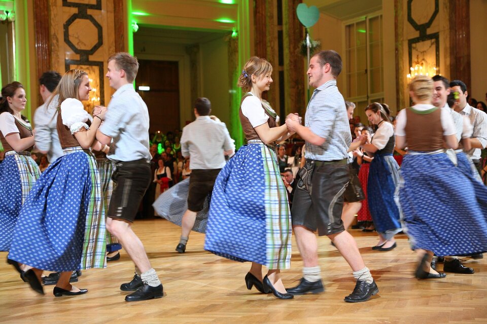 Eine Gruppe von Menschen tanzt in traditioneller Tracht auf einem festlichen Ball. Die Atmosphäre ist lebhaft und fröhlich, mit vielen Gästen im Hintergrund.