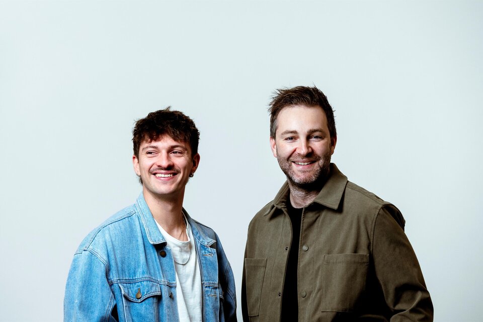 Zwei Männer stehen freundlich nebeneinander vor einem hellen Hintergrund. Sie lächeln und tragen legere Kleidung. | © @mario.wallner