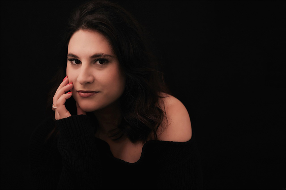 Eine Frau mit dunklen, lockigen Haaren sitzt vor einem schwarzen Hintergrund. Sie trägt einen schwarzen Pullover und schaut nachdenklich in die Kamera. | © Simon Petek