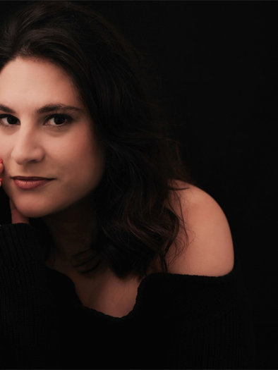 A woman with dark, curly hair sits in front of a black background. She is wearing a black sweater and is looking thoughtfully at the camera. | © Simon Petek