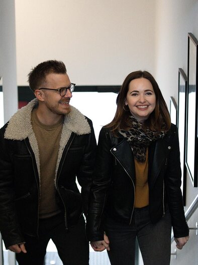 A couple is smiling as they walk up a staircase. They are wearing fashionable jackets and seem happy together. | © achromaticphotography