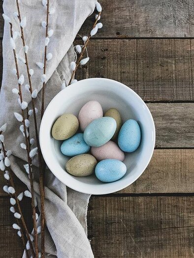 A white bowl with colorful eggs is on a wooden table. Next to it lies a beige cloth and some pussy willow branches. | © Pixabay