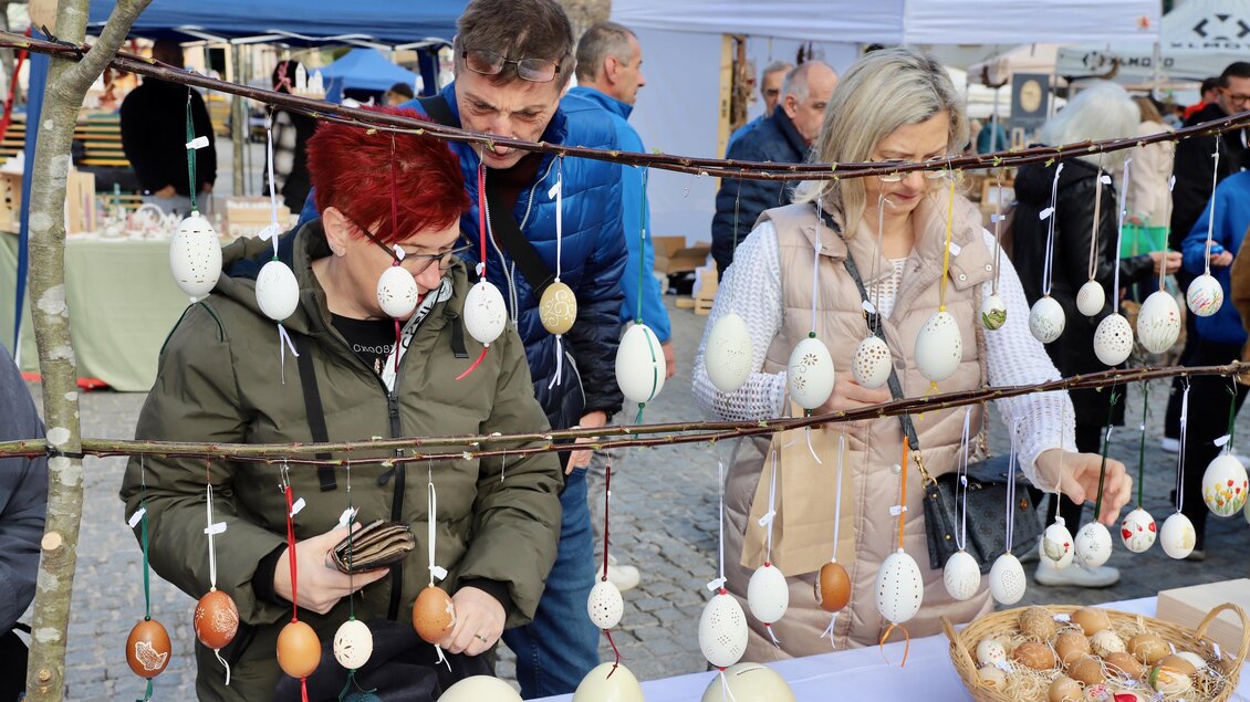 Ein Marktstand mit kunstvoll bemalten Eiern, die an einem Ast hängen. Zwei Personen betrachten die Eier, während eine dritte Person im Hintergrund zuschaut. | © Oststeiermark Tourismus