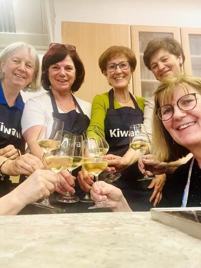A group of seven women is celebrating with champagne glasses in a kitchen. They all wear aprons and smile happily at the camera. | ©  Lipp Eva Maria