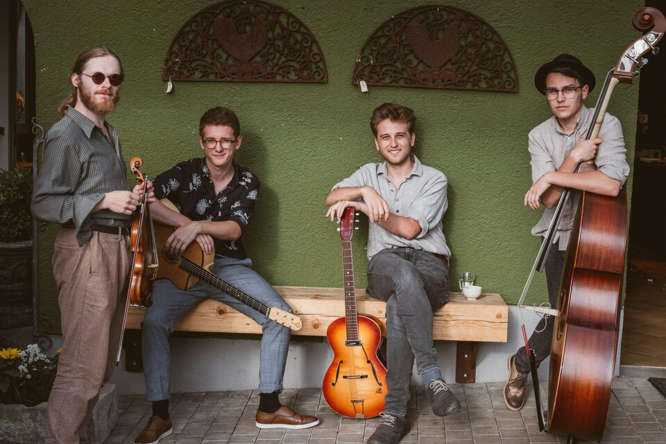 Eine vierköpfige Band sitzt auf einer Bank und posiert. Die Musiker halten verschiedene Instrumente, darunter eine Gitarre und eine Violine. | © Paganin Soatnquartett