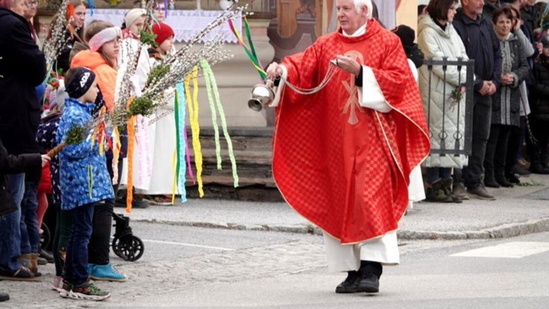 Ein Priester in einem roten Gewand steht auf einer Straße und läutet eine Glocke. Um ihn herum stehen Menschen, darunter Kinder mit bunten Zweigen und Blumen. | © Mag. Herbert Ribul