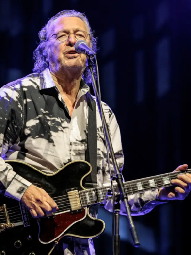 A musician sings into a microphone and plays guitar. He is wearing a striking shirt and has long hair. | © ©Alexander Mueller