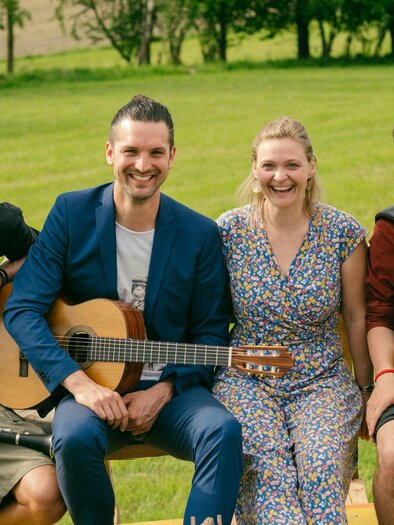 A young man stands in a green meadow surrounded by trees. In the background, gentle hills and a cloudy sky can be seen. | © Manuel Hauer