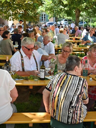 A lively outdoor restaurant with many guests sitting at long tables. The people enjoy food and drinks under shady trees. | © Pfarre Gamlitz