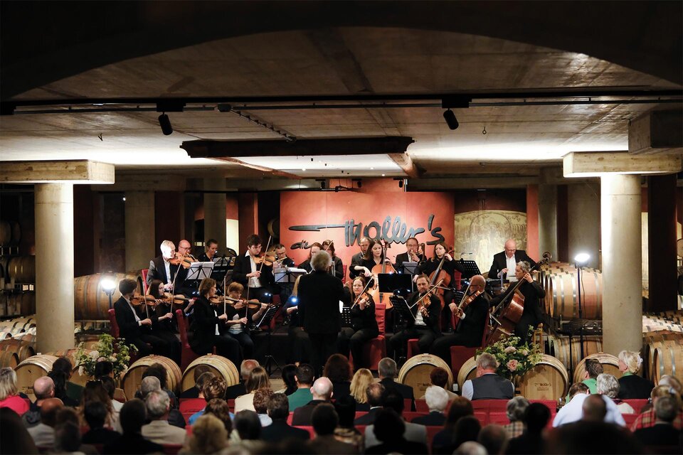 Ein Orchester spielt live in einem stilvollen Raum mit Fässern im Hintergrund. Das Publikum sitzt gespannt und lauscht der Musik. | © Weinschloss Thaller