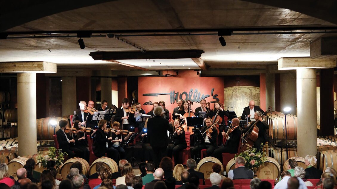 Ein Orchester spielt live in einem stilvollen Raum mit Fässern im Hintergrund. Das Publikum sitzt gespannt und lauscht der Musik. | © Weinschloss Thaller