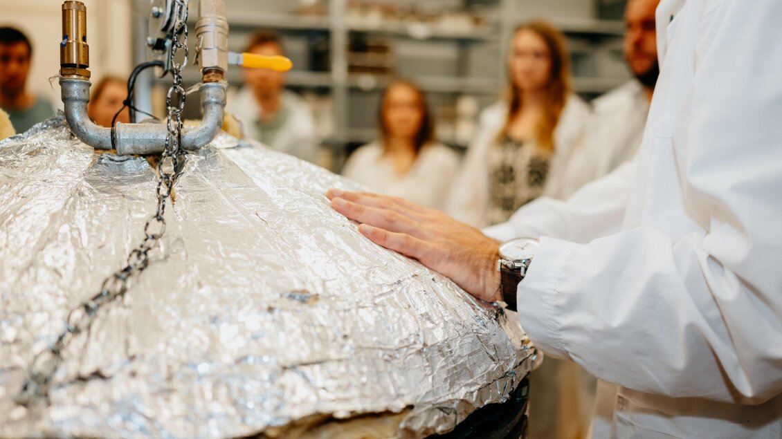 Eine Gruppe von Menschen in Laborkleidung beobachtet eine mit Folie bedeckte Apparatur. Jemand legt seine Hand auf das Gerät, während eine wissenschaftliche Demonstration stattfindet.